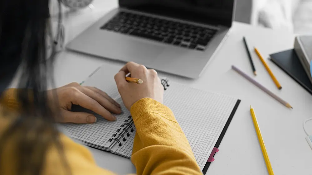Persona escribiendo en un cuaderno junto a un computador.